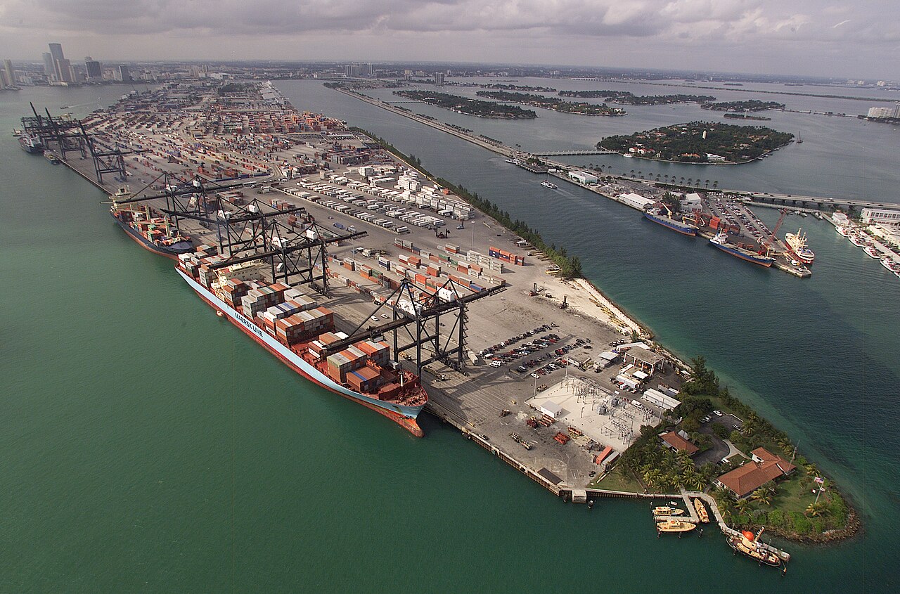 Large container ship being unloaded by cranes at Port Miami, with the downtown Miami skyline in the background.